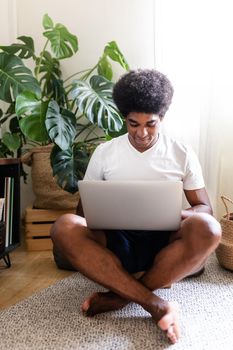Calm and serene young african american man sitting cross-legged on the floor writing an email in his laptop. Vertical image. At home concept. Technology concept.