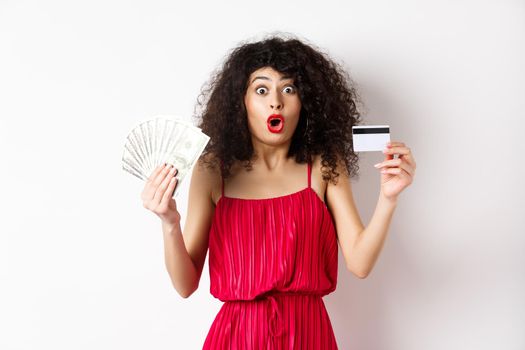 Impressed lady with curly hair and trendy red dress, saying wow, showing plastic credit card with cash, standing on white background.