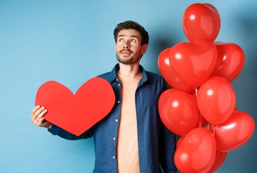 Valentines day and love concept. Man dreaming of soulmate, holding big red heart cutout and balloons, standing over blue background.