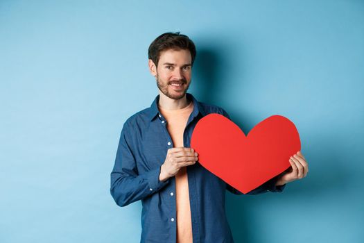 Happy man showing valentines heart and smiling, make romantic gift on lovers day, standing over blue background.