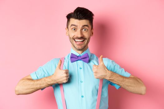 Happy young man showing thumbs up and smiling, saying yes, agree with something cool, recommending excellent deal, standing upbeat on pink background.