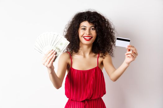 Shopping. Lucky woman in elegant dress, showing money and plastic credit card, smiling pleased, wearing makeup, white background.