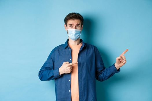 Covid and healthcare concept. Worried young man in medical mask raising eyebrow, pointing finger right with doubtful and nervous face, standing on blue background.