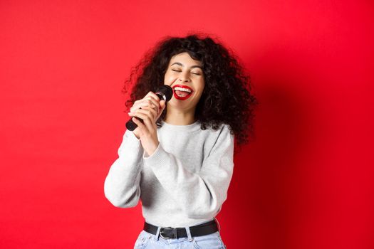 Hobbies and leisure concept. Happy woman singing song in microphone, having fun at karaoke with mic, standing on red background.