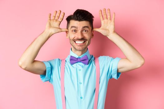 Silly and childish guy in suspenders and bow-tie, mocking someone, fooling around and making funny gestures, standing on pink background.