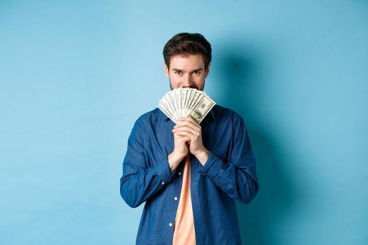Young man hiding smiling face behind money and looking determined at camera, standing on blue background.