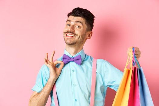 Confident smiling guy adjusting his bow-tie and showing gifts in shopping bags, looking happy, standing over pink background.