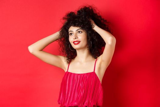 Carefree female model playing with hair, looking dreamy and relaxed at logo, smiling sensual, standing on red background.