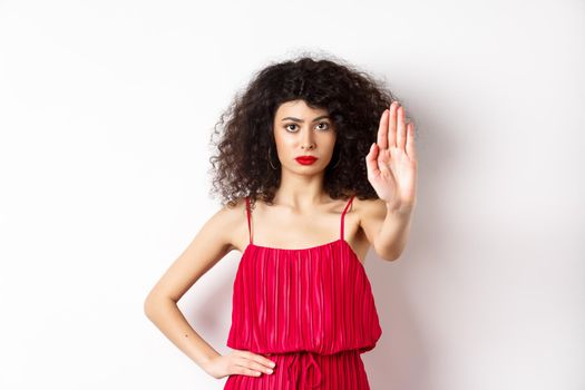 Serious and confident woman in red dress and makeup stretch out hand, tell to stop, prohibit and forbid something, standing over white background.