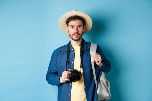 Handsome young tourist in summer hat, backpacking and travelling, holding photo camera, standing on blue background.