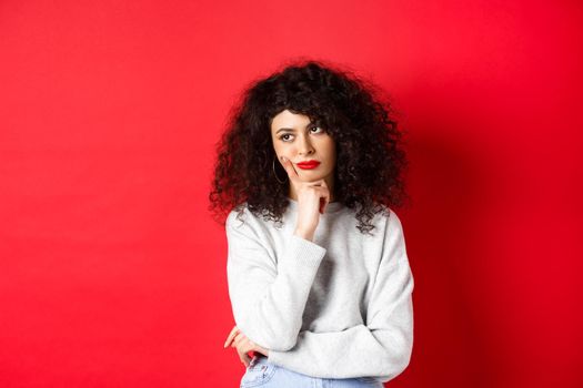 Grumpy young woman with curly hair, looking annoyed or bored at empty space, standing pensive and sad on red background.