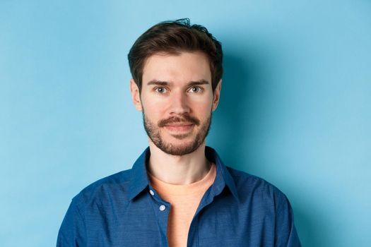 Close-up of young caucasian guy with beard smiling at looking happy at camera, standing on blue background.