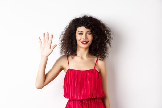 Cheerful young woman with makeup and red dress, showing five fingers and smiling, standing over white background. Copy space