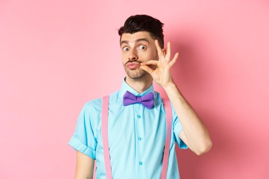 Funny young man touching his french moustache and pucker lips, looking silly at camera, standing in bow-tie and suspenders on pink background.