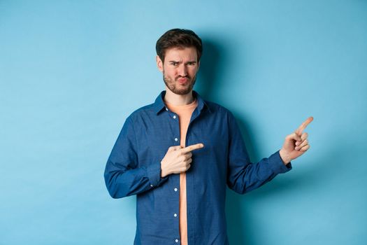 Disappointed young man frowning and pointing fingers right at empty space, looking upset and displeased, complaining, standing on blue background.
