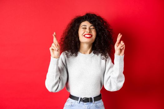 Hopeful and positive woman with red lips and curly hair, cross fingers for good luck and making wish, praying for dream come true, smiling excited, red background.
