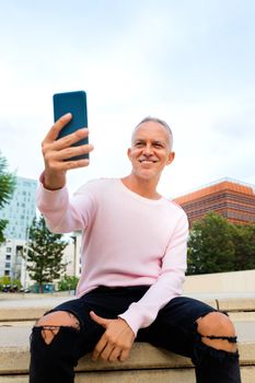 Caucasian adult man with pink sweater taking a selfie outdoors using smartphone. Vertical image. Social media and technology concept.