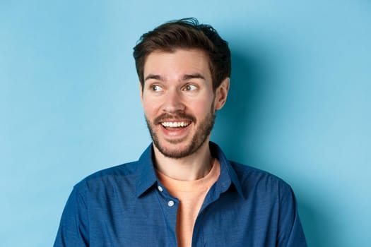Close-up of cheerful bearded guy looking left at empty space and smiling, standing happy on blue background.