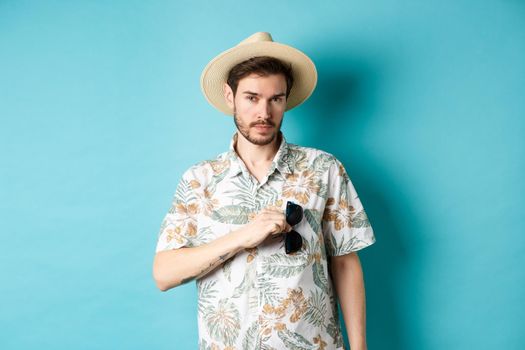 Handsome tourist in hawaiian shirt and summer hat, put sunglasses in pocket, going on vacation, standing against blue background.