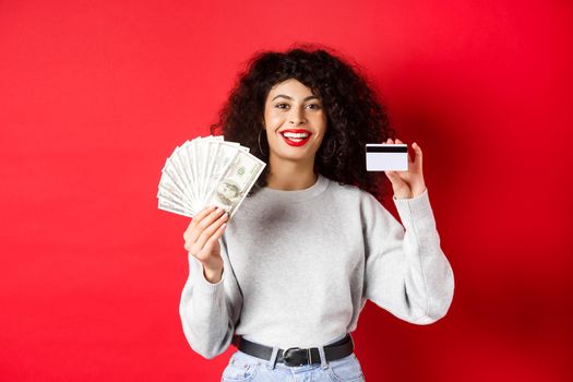 Portrait of stylish young woman with curly hair, showing money in cash and plastic credit card, red background.
