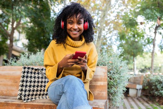 Young African American woman wearing headphones using cellphone sits on park bench looking at camera. Copy space. Lifestyle and technology concepts.