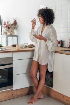 Young African American woman drinking water and using cellphone in the kitchen. Vertical image. Lifestyle concept.