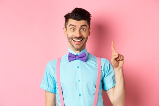 Cheerful caucasian man pitching an idea, raising finger in eureka gesture and smiling, suggesting solution, standing on pink background.