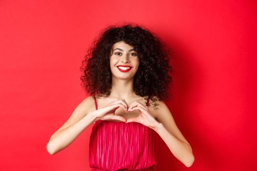 Valentines day. Romantic girl with curly hairstyle in evening dress, smiling and showing heart sign, say I love you on lovers holiday, standing over red background.