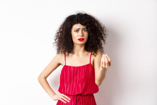 Confused woman with curly hair, wearing red dress, frowning and shaking hand, cant understand something, looking hesitant, standing on white background.