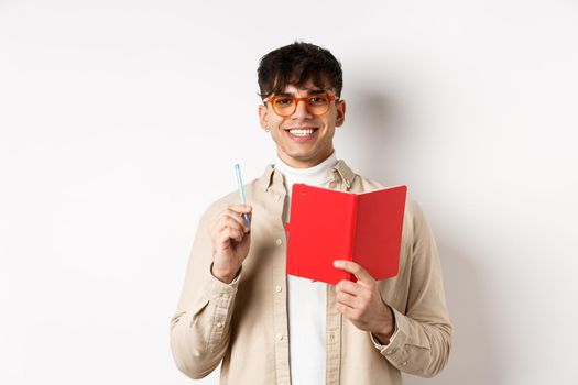 Happy person in glasses writing in journal, holding pen and diary, smiling at camera, plan a schedule, standing with planner on white background.