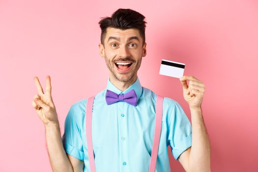 Shopping concept. Smiling caucasian guy in bow-tie showing plastic credit card and peace, victory sign, standing happy on pink background.