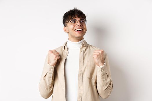 Lucky young man say yes with relieved face, smiling and making fist pump, triumphing of success, winning prize, standing on white background after achieving goal.