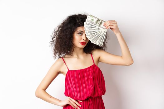 Elegant curly-haired woman in red dress, showing fan of money and looking confident. Rich girl holding dollar bills, white background.