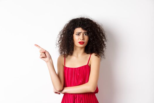 Arrogant young woman with curly hair, wearing red dress, frowning and complaining, pointing finger left at promo, standing over white background.