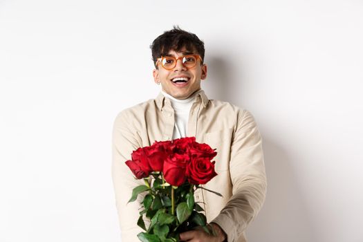 Man in love holding red roses and looking tenderly at camera, staring at lover with happy face, celebrating Valentines day with girlfriend, standing over white background.