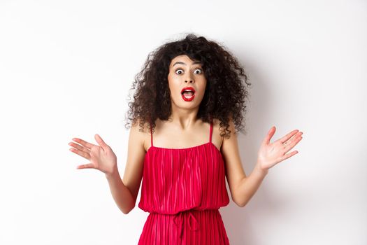 Excited woman with curly hair, talking with amazed face, explain big news, shaking hands and look astonished at camera, white background.