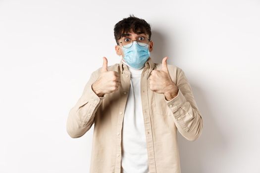 Covid-19, health and real people concept. Cheerful man in glasses and sterile face mask, showing thumbs up in approval, praise and compliment product, standing on white background.