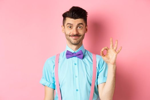 Smiling caucasian man in bow-tie and suspenders showing all okay gesture, recommending something cool, praising promo on pink background.