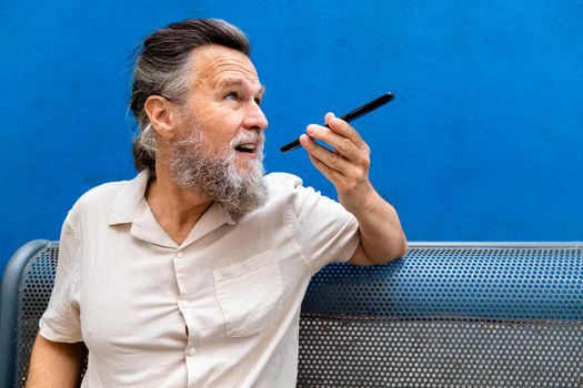 Mature caucasian man with beard sitting on a bench sending voice message using mobile phone. Copy space. Technology concept. Lifestyle.