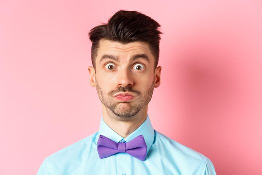 Close-up of confused caucasian guy holding breath, pouting and staring shocked at camera, wearing festive clothes and bow-tie, pink background.