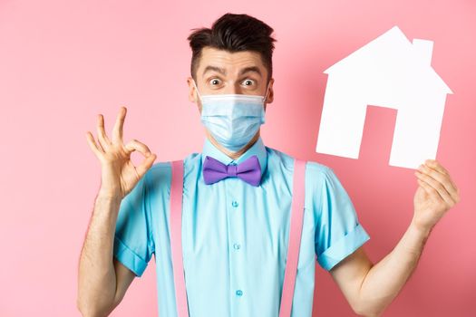 Covid, pandemic and real estate concept. Excited young man in medical mask showing OK sign and paper house cutout, recommending agency, standing over pink background.