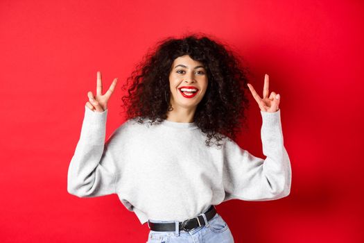 Stylish young woman with curly hairstyle, smiling happy and showing peace signs, standing in casual sweatshirt on red background.
