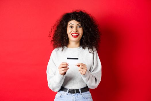 Excited smiling woman showing plastic credit card, standing in casual clothes on red background.