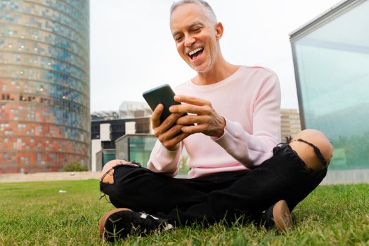 Smiling caucasian man sitting on grass in city public park looking at cellphone. Social media and technology concept.