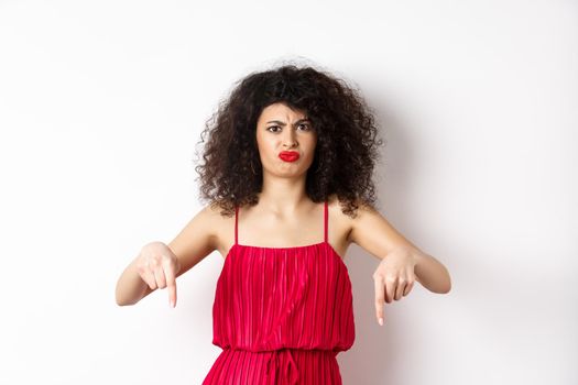 Disappointed caucasian woman in red dress complaining on spoiled date, frowning and grimacing, standing over white background.