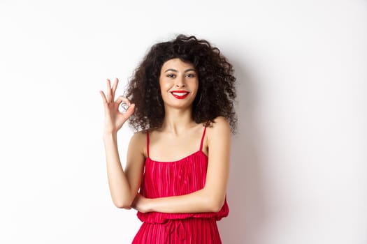 Elegant woman in romantic red dress and makeup, showing okay sign and smiling, recommending good thing, approve and agree, standing satisfied on white background.