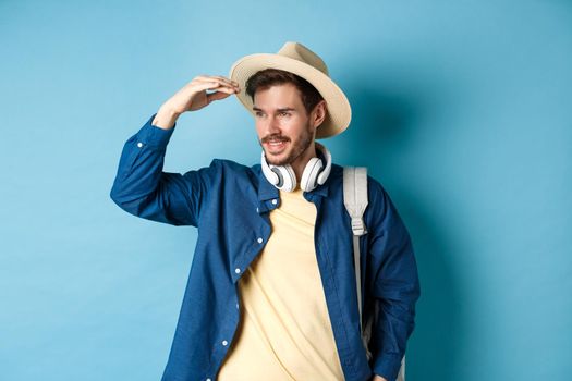 Handsome tourist in straw hat, covering face from sun and looking at distance aside, smiling pleased, travelling on summer vacation, standing over blue background.