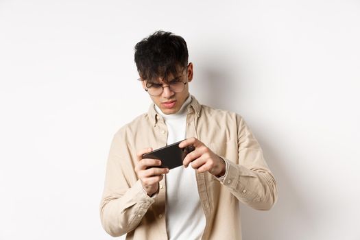 Portrait of handsome young man playing video game on mobile phone, tilt smartphone to play racing, standing on white background.