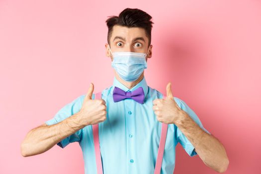 Coronavirus, healthcare and quarantine concept. Satisfied guy in medical mask showing thumbs up in approval, praising excellent choice, pink background.