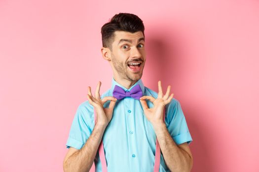 Classy handsome man fixing bow-tie on neck and smiling, getting ready for date or party, standing on pink background.